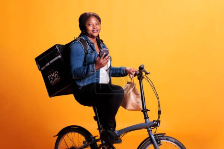 Téléchargez les photos : African american restaurant courier looking at client adreess on smartphone before start delivering fast food order to client. Pizzeria employee bringing takeaway meal using bike as transportation - en image libre de droit