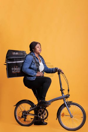 Foto de Restaurant courier holding thermal backpack delivering takeaway food order to client during lunch time, standing over yellow background. Pizzeria delivery worker riding bike to deliver fast food meal - Imagen libre de derechos