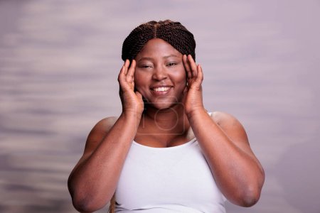 Foto de Happy curvy woman with natural makeup touching face portrait. Carefree body positive african american beauty model putting hands on fresh healthy skin and doing skincare procedures - Imagen libre de derechos