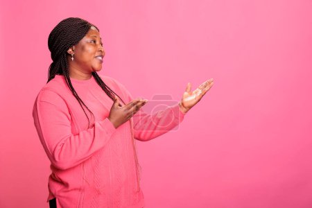 Foto de Smiling confident woman pointing to side with palm while advertising promotional product in studio. Cheerful young adut showing promotion standing over pink background, advertise gesture - Imagen libre de derechos