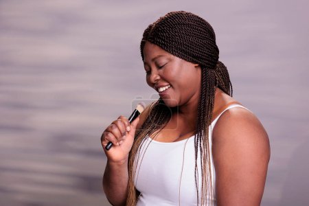 Téléchargez les photos : Femme afro-américaine chantant une chanson au pinceau de maquillage tout en appliquant des cosmétiques décoratifs et en écoutant de la musique. Dame courbée faisant maquillage et en utilisant l'outil de cosmétologie comme micro - en image libre de droit