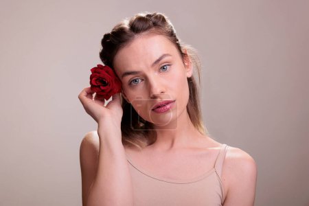 Photo for Attractive caucasian woman putting red rose in blonde hair face portrait. Beautiful young lady holding bright flower bud behind ear and looking at camera with romantic glance - Royalty Free Image