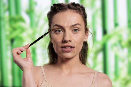 Téléchargez les photos : Young beautiful woman doing eyes makeup with brush and looking at camera. Attractive beauty model using natural green decorative cosmetics and applying eyeshadows portrait - en image libre de droit