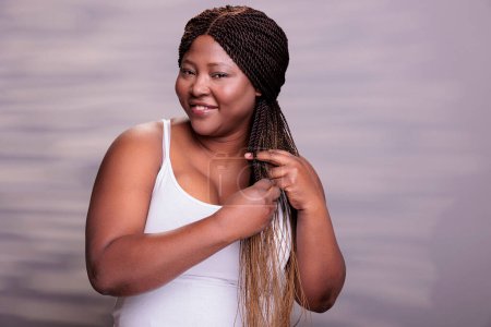Photo for Plus size african american model making braids hairstyle and looking at camera. Beautiful happy body positive curvy woman smiling with cheerful facial expression braiding hair portrait - Royalty Free Image