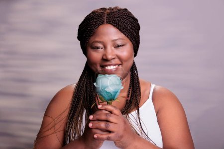 Photo for Smiling happy young curvy lady holding flower portrait, showing self care and body positivism concept. Attractive carefree african american lady posing in studio with blue rose - Royalty Free Image