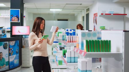 Foto de Joven examinando cajas de medicamentos en estantes en farmacia, buscando medicamentos recetados y productos farmacéuticos. Mujer que revisa suplementos médicos yvitaminas d. - Imagen libre de derechos