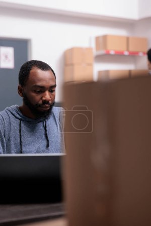 Foto de Storehouse manager checking shipment logistics before start preparing packages, putting customer order in carton boxes. African american supervisor working in warehouse delivery department - Imagen libre de derechos