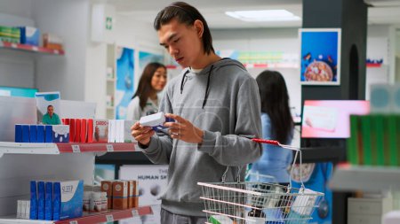 Foto de Male customer looking at medicaments boxes in drugstore, searching for medical treatment and vitamins. Asian adult analyzing packages of pharmaceutics before buying supplements or pills. - Imagen libre de derechos