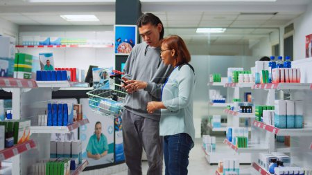 Foto de Asian worker reading prescription on smartphone to give medicine to client, explaining healthcare treatment in pharmacy store. Drugstore consultant helping young man with products and pills. - Imagen libre de derechos