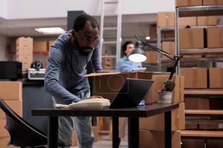 Téléchargez les photos : African american storehouse manager checking stock cargo on laptop computer while preparing packages, putting clients orders in carton boxes. Team working in delivery department in warehouse - en image libre de droit