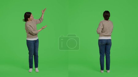 Téléchargez les photos : Femme souriante décontractée pointant vers l'icône de présentation en studio, faisant de la publicité et montrant l'exemple à la caméra. Personne posant sur fond d'écran vert isolé, fille confiante faisant de la publicité. - en image libre de droit