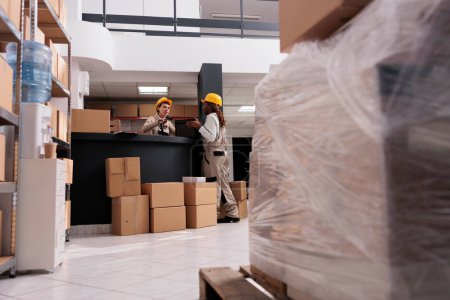 Foto de Retail storehouse operative managing product receiving and scanning suticase at desk. Two diverse employees wearing protective overalls working in warehouse full of cardboard boxes parcels - Imagen libre de derechos