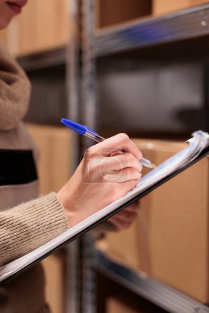 Téléchargez les photos : Post office warehouse employee searching parcel, checking postal form. Young caucasian woman supervising cardboard boxes in mail sorting center and writing on clipboard close up - en image libre de droit