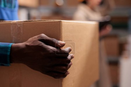 Téléchargez les photos : Shipping service warehouse african american man worker holding received parcel in hands. Delivery company employee lifting heavy cardboard box in storehouse with close view on arm - en image libre de droit
