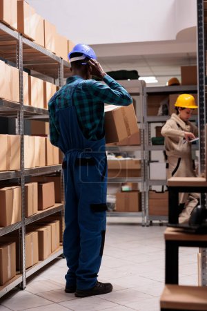 Téléchargez les photos : Delivery service worker holding cardboard box, preparing parcel for transportation in storage. Shipping business company warehouse operative wearing protective unifrom carrying carton - en image libre de droit
