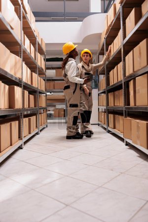 Photo for Freight distribution center employees scanning boxes before transportation. Two woman warehouse employees wearing protective helmets and overalls talking, standing near shelf full of cartons - Royalty Free Image