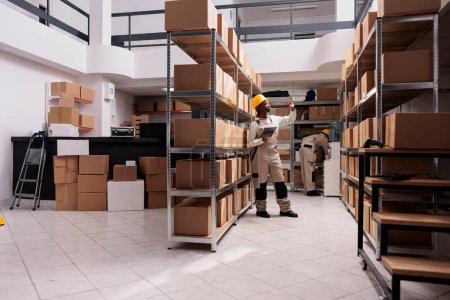 Téléchargez les photos : Shipping company worker standing in warehouse aisle near cardboard boxes shelf, using digital tablet and searching customer parcel. Delivery service storage employee looking at packages on metal racks - en image libre de droit