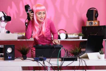 Téléchargez les photos : Asian musician standing at dj table enjoying to perform electronic music in front of crowd, using professional turntables in club at night. Performer with pink hair playing techno song - en image libre de droit