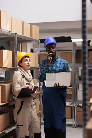 Téléchargez les photos : Warehouse workers checking factory goods supply and monitoring dispatching operations on laptop. Caucasian and african american storehouse colleagues standing near cardboard boxes shelf - en image libre de droit