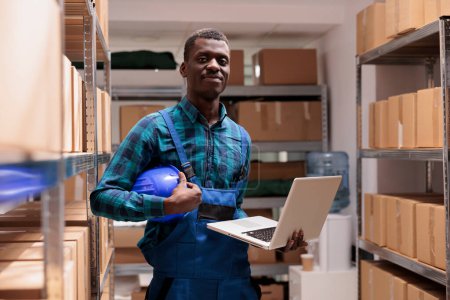 Foto de Warehouse employee doing inventory management using laptop, checking parcels before transportation and looking at camera. Smiling african american storehouse worker portrait - Imagen libre de derechos