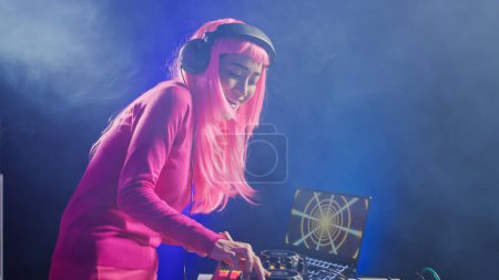 Téléchargez les photos : Smiling artist standing at dj table playing electronic music at professional mixer console in studio over pink background. Asian musician performing techno sound, having fun in club at night time - en image libre de droit