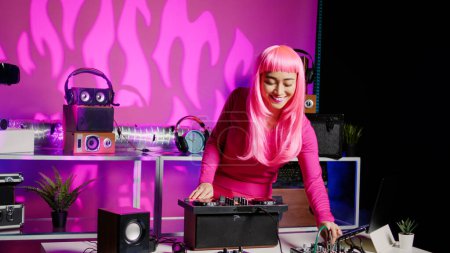 Foto de Smiling musician working as dj playing at mixer console, mixing techno sound with eletronic using audio equipment. Performer with pink hair having fun performing in club at night time - Imagen libre de derechos