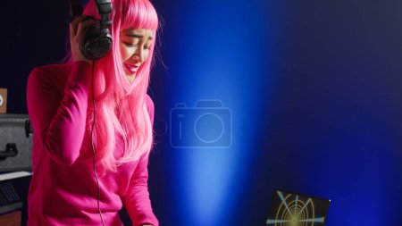Foto de Dj with pink hair playing eletronic song at professional mixer console, listening music into headset. Asian perfomer enjoying mixing sounds, dancing with fans during night time in club. - Imagen libre de derechos