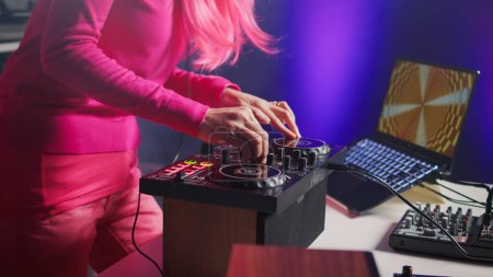 Foto de Musician mixing electronic sound with techno, enjoying party with fans, standing at dj table performing concert in club at night. Asian artist with pink hair performing music using turntables - Imagen libre de derechos