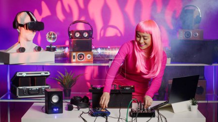 Téléchargez les photos : Cheerful performer dancing and interacting with fans in club at nightime, playing techno remix at professional mixer console. Artist with pink hair mixing electronic sounds using audio equipment - en image libre de droit