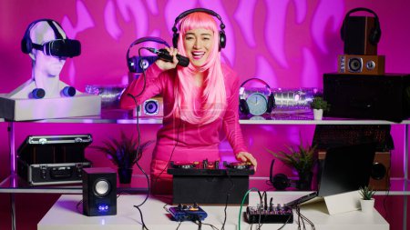 Téléchargez les photos : Dj woman putting headphones before start mixing music using audio equipment during techno party in nightclub. Asian performer with pink hair creating musical performance with mixer console - en image libre de droit