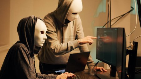 Téléchargez les photos : Mysterious spies working on phishing and cryptojacking, trying to break into government server and steal valuable data. Team of hackers with anonymous masks hacking online system. - en image libre de droit
