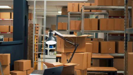 Photo for Empty office space in storage room warehouse filled with cardboard packages, desk with laptop and inventory or logistics notes. Storespace with stacks of merchandise in carton boxes. - Royalty Free Image