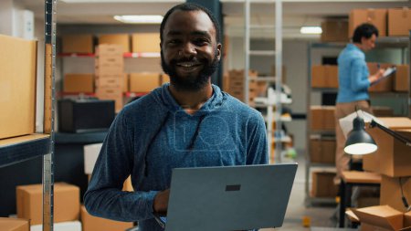 Photo for Warehouse employee using laptop to write products information, analyzing racks filled with carton packages for stock inventory. Young man planning logistics and merchandise management. - Royalty Free Image