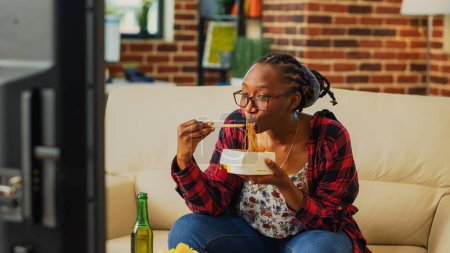 Téléchargez les photos : Cheerful girl using chopsticks to eat noodles, laughing at favorite comedy movie on television. Young adult eating asian food in takeaway box and drinking alcohol, having fun at tv. - en image libre de droit