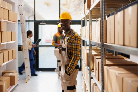 Foto de African american smiling warehouse worker standing near cardboard boxes shelf in stockroom portrait. Young storehouse man employee wearing protective helmet and overall and looking at camera - Imagen libre de derechos