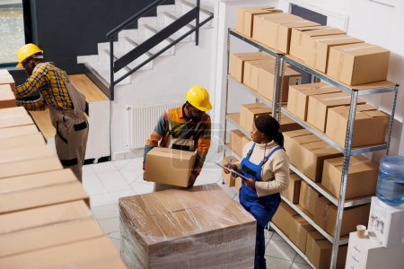Foto de Package handler holding parcel and talking with supervisor in warehouse. African american logistics manager inspecting freight transportation and checking cardboard box in storehouse - Imagen libre de derechos