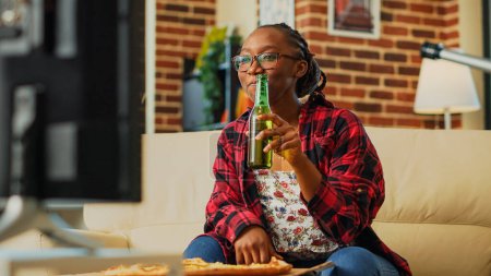 Photo for Smiling woman eating pizza slice in living room, enjoying comedy movie on television. Young person feeling happy drinking bottle of beer and having dinner from takeaway delivery. Handheld shot. - Royalty Free Image