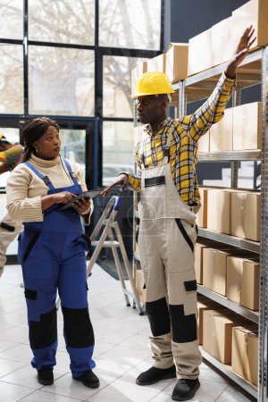 Photo for African american warehouse worker explaining supervisor packages storage system, showing on parcels shelf and talking. Storehouse manager listening to colleague and holding digital tablet - Royalty Free Image