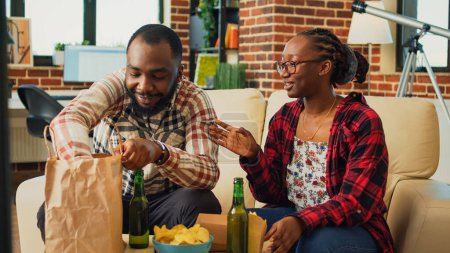 Téléchargez les photos : African american couple eating burgers with fries at home, having fun watching movie on tv and drinking beer. Boyfriend and girlfriend serving fast food meal from takeaway restaurant. - en image libre de droit