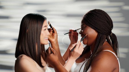 Photo for Diverse women playing with make up brushes in studio, giving eachother makeovers and having fun as friends. Young beautiful girls enjoying skincare and body positivity campaign. - Royalty Free Image