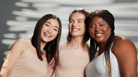 Téléchargez les photos : Multiethnic group of models posing for body positivity ad in studio, having fun with friends advertising skincare products. Cheerful women laughing on camera, different skintones and body types. - en image libre de droit