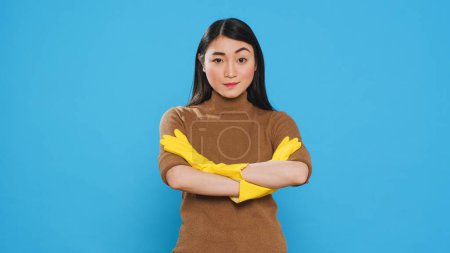 Photo for Asian professional maid smiling at camera after finishing house cleaning, standing in studio over blue background. Housekepper was skilled in the art of housekeeping, ensuring every surface was clean - Royalty Free Image