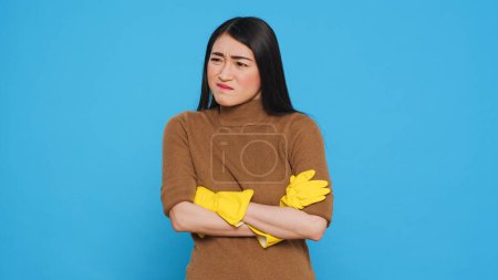 Photo for Nervous professional cleaner with protective yellow gloves having problem with house cleaning, standing with arm crossed in studio over blue background. Housekepper having negative expression - Royalty Free Image