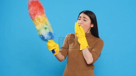Téléchargez les photos : Tired professional maid cleaning furniture dust using colorful duster during cleaning session in studio. Asian housekepper committed to maintaining high standards of home cleanliness - en image libre de droit