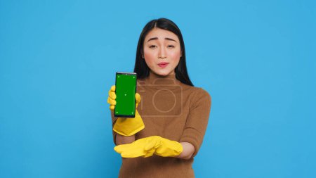 Téléchargez les photos : Asian homemaker holding chroma key mock up mobile phone with greenscreen display while talking with remote client during videocall meeting. Cheerful maid was skilled in all aspects of housework - en image libre de droit