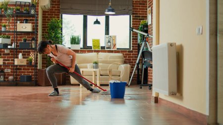 Foto de Caucasian wife cleaning apartment floors with mop, listening to music and doing chores. Cheerful woman dancing and washing dirt, doing housework in living room. Spring cleaning. - Imagen libre de derechos