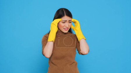 Téléchargez les photos : Overwhelmed housekeeper being tired after cleaning all house using variety of chemical detergents, standing in studio over blue background. Housewives relied on the cleaning lady services - en image libre de droit