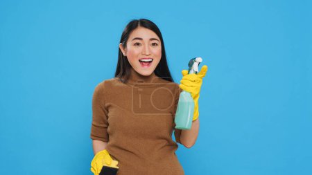 Photo for Professional housekeeper being amazed about how good is detergent spray while cleaning house for her clients. Asian maid committed to maintaining the highest standards of cleanliness and hygiene - Royalty Free Image