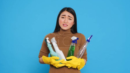 Téléchargez les photos : Overwhelmed tired housekeeper wearing gloves while holding multiple sanitary cleanliness products while cleaning house for clients. Stressed maid ensuring that everything was kept clean and organized. - en image libre de droit
