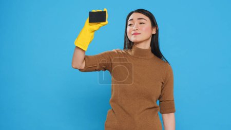 Téléchargez les photos : Smiling maid using sponge and professional cleaning detergent while cleaning customer house, using cleanliness equipment. Cleaner wore gloves as part of her attire while doing the housework. - en image libre de droit
